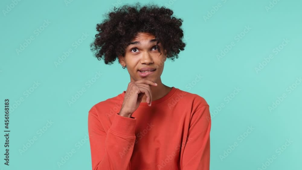Young curly african american man 20s wears azure t-shirt looks around thinks put hand prop up on chin comes up with ideas raised finger isolated on plain pastel light blue background studio portrait Young curly african american man 20s wears azure t-shirt looks around thinks put hand prop up on chin comes up with ideas raised finger isolated on plain pastel light blue background studio portrait