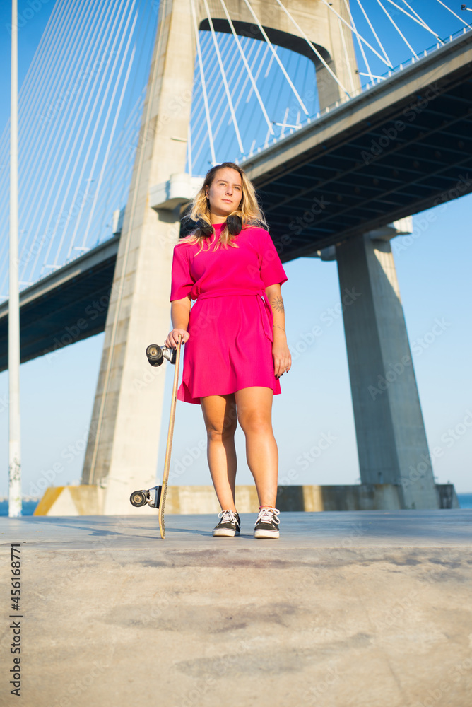 Portrait of pretty young woman holding longboard. Beautiful girl in headphones and dress with skateboard standing near bridge. Sport, hobby, active lifestyle concept