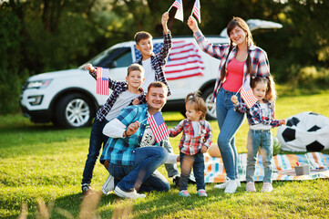 large american family spending time together. With USA flags against big suv car outdoor. America holiday. Four kids.