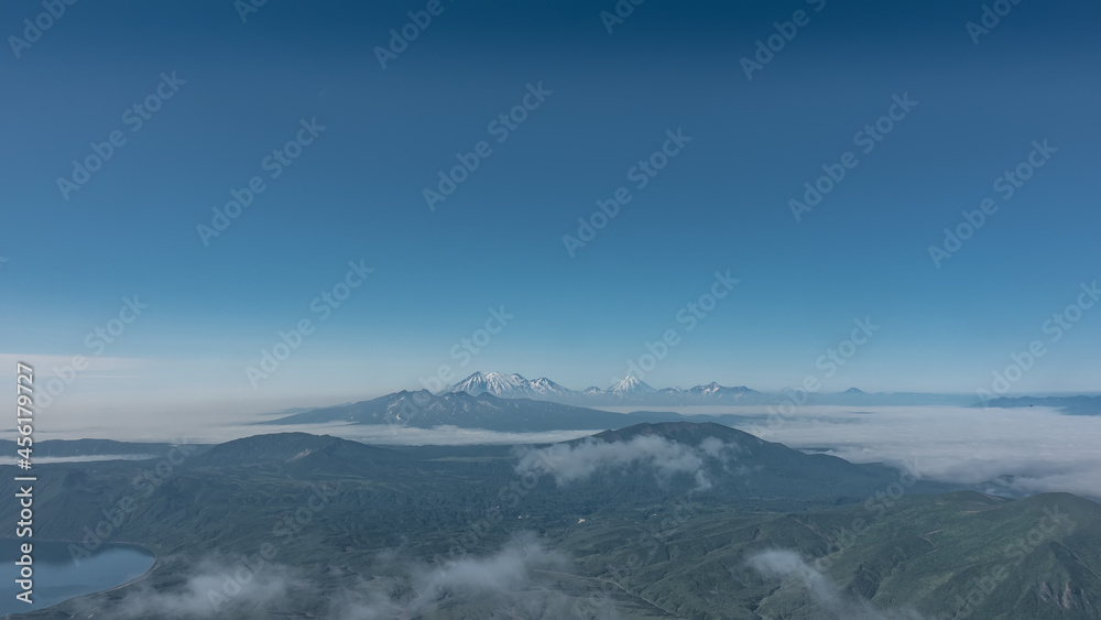 Kamchatka landscape, view from a height. Clouds float over the green mountains and the lake. In the distance, against the blue sky, a ridge of conical volcanoes with snow-covered slopes