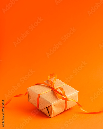 Close up of brown gift box with a orange satin ribbon bow on orange blurred background with copy space. Holiday autumn concept