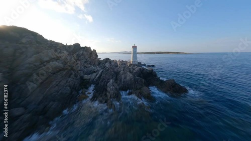 FPV video, view from above, stunning aerial view from an FPV drone flying at high speed over a rocky coastline with a lighthouse illuminated during a dramatic sunset. Faro di Capo Ferro, Sardinia.