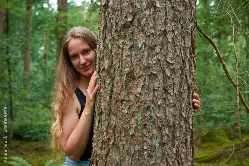 A blonde Caucasian lady with long hair looking from behind a tree trunk and smiling A blonde Caucasian lady with long hair looking from behind a tree trunk and smiling