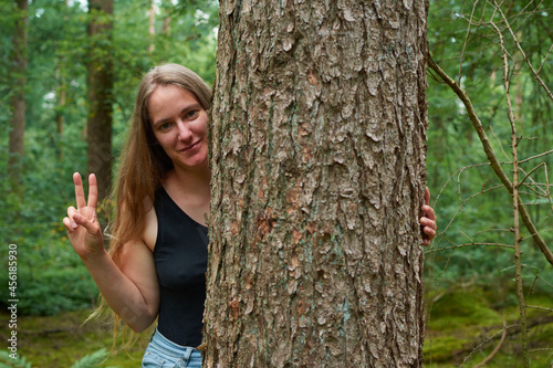 A blonde Caucasian lady with long hair looking from behind a tree trunk showing the peace gesture