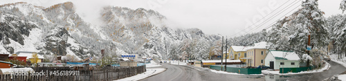 Chemal, Altai Republic, Russia - 15 October 2020: Panoramic view. Russian rural village Chemal in the snow. The main road against the background of the Altai mountains, forests and clouds.