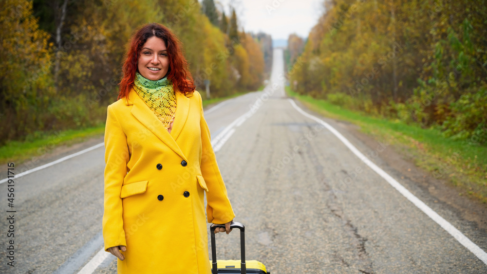 Beautiful red-haired woman in a yellow coat walks with a yellow suitcase along the highway in autumn. Beautiful red-haired woman in a yellow coat walks with a yellow suitcase along the highway in autumn.