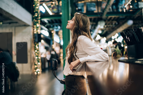 Young female customer in white shirt thinking about restaurant business standing near bar indoors, contemplative hipster girl with curly brunette hair pondering on idea for spending leisure weekend