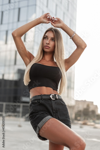 Fashionable beautiful model woman in black stylish t-shirt and denim shorts with belt posing in the city