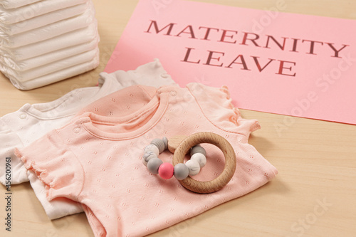 Baby clothes, toy, diapers and note with text Maternity Leave on wooden table