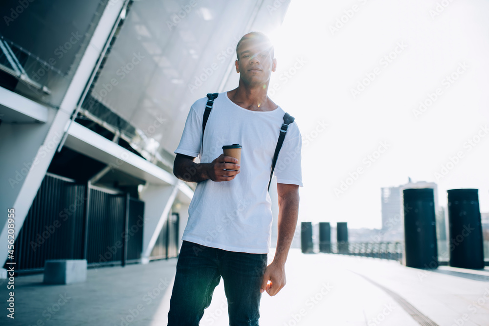 Serious young man with paper cup of coffee walking on street