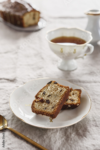 Slice of Pound Loaf Cake with raisins on white porcelain plate and cup of tea on greige linen tablecloth. Selective focus