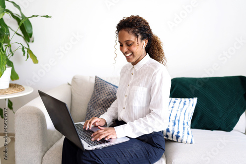 Black woman with long curly hair with ear buds earphones using laptop wotking sitting on sofa wearing smart casual clothing