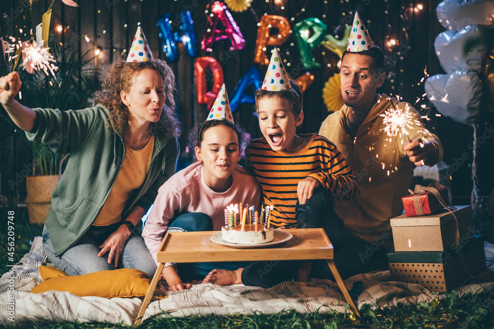 Happy parents with sparklers and children trying to blow burning candles on delicious birthday cake celebrate holiday in cottage yard in evening