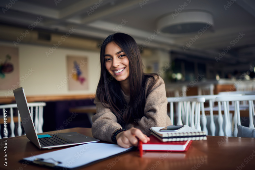 Portrait of joyful Middle Eastern female student with digital netbook device and education equipment smiling at camera during coworking e learning time, happy freelancer with laptop computer laughing Portrait of joyful Middle Eastern female student with digital netbook device and education equipment smiling at camera during coworking e learning time, happy freelancer with laptop computer laughing