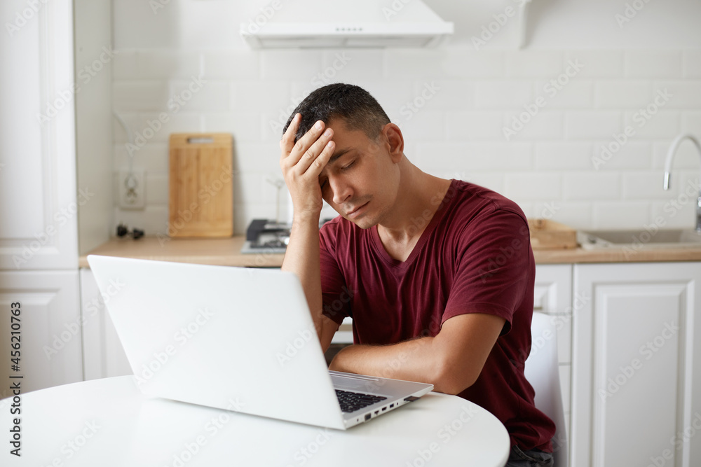 Portrait of tired man wearing maroon casual style t shirt sitting at table against kitchen set, keeps eyes closed, having headache, working online long hours, being exhausted. Portrait of tired man wearing maroon casual style t shirt sitting at table against kitchen set, keeps eyes closed, having headache, working online long hours, being exhausted.