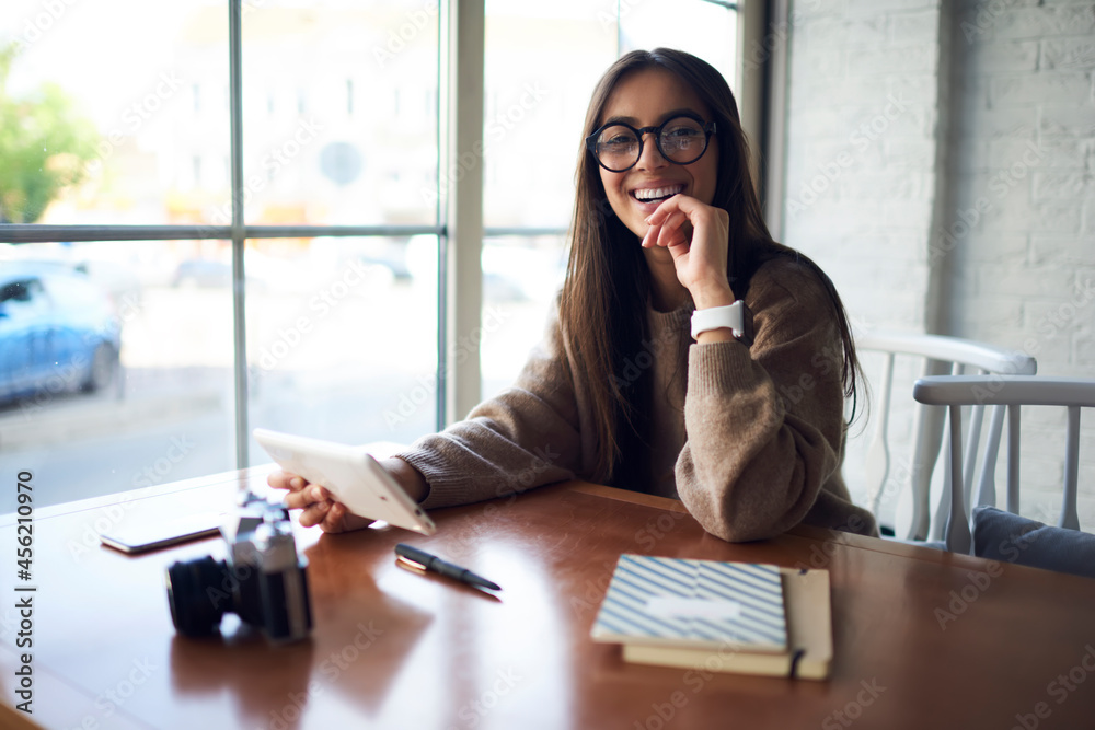Portrait of cheerful hipster girl in stylish spectacles for vision protection smiling at camera during free time in cafe interior, happy Middle Eastern female blogger with touch pad technology - obrazy, fototapety, plakaty 