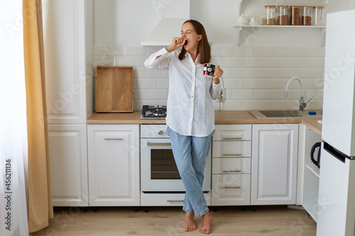 Foto Full length portrait of sleepy female drinking hot coffee in the morning, standing with cup in hands and yawning, needs energy, wearing white shirt and jeans