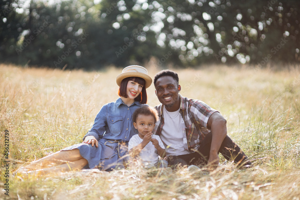 Portrait of beautiful and happy family of three having summer pinic on nature. Cute little boy sitting in hugs with multicultural parents and eating sweet grape. Portrait of beautiful and happy family of three having summer pinic on nature. Cute little boy sitting in hugs with multicultural parents and eating sweet grape.