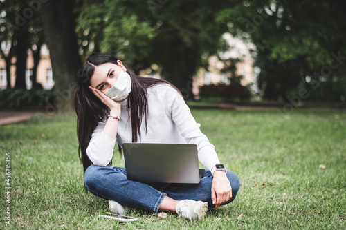 A young girl in a protective mask works in the park on a laptop and with a phone