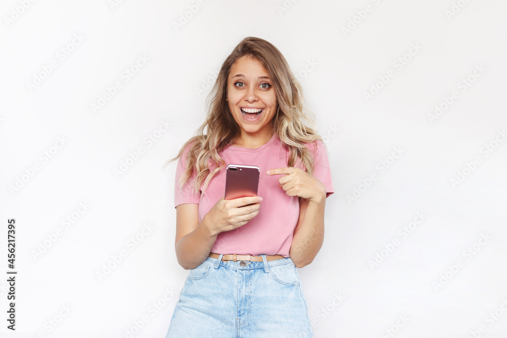 A young pretty caucasian impressed excited smiling blonde woman in a pink t-shirt holds mobile phone in her hand and points at the screen with her finger isolated on a white background A young pretty caucasian impressed excited smiling blonde woman in a pink t-shirt holds mobile phone in her hand and points at the screen with her finger isolated on a white background
