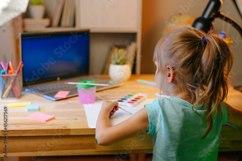 Little girl elementary student drawing studying online from home using laptop sitting at the desk