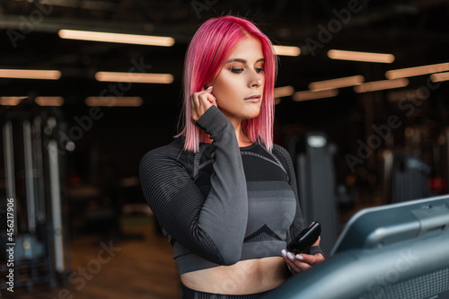 Young beautiful fitness woman with pink hair with black wireless headphones listening to music and running in the gym