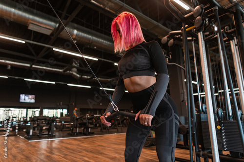 Sports beautiful young girl model with a slender body and pink hair in a fashionable black tracksuit working out in the gym