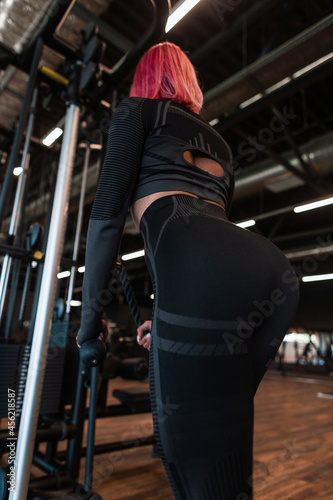 Pretty young fitness woman with a slender beautiful body in black sportswear works out on the exercise machine in the gym, back view