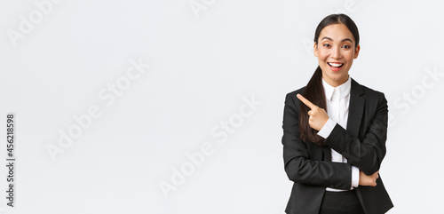 Excited attractive asian female saleswoman, real estate agent in suit suggesting perfect house, standing in suit and pointing finger left. Businesswoman making announcement, show banner photo