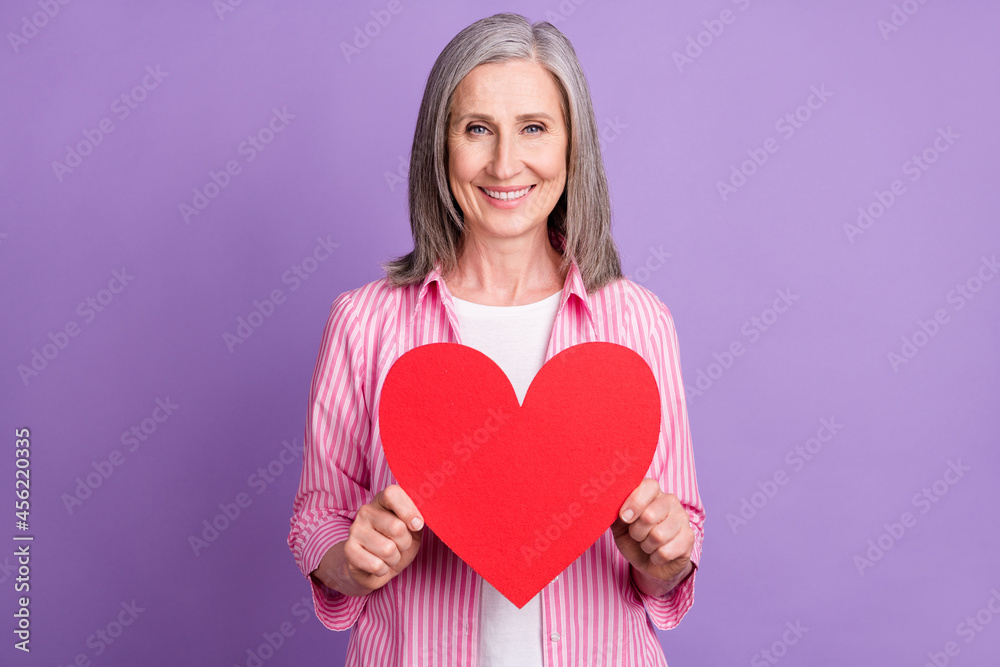Photo of cheerful mature woman happy positive smile hold red heart romance isolated over violet color background Photo of cheerful mature woman happy positive smile hold red heart romance isolated over violet color background