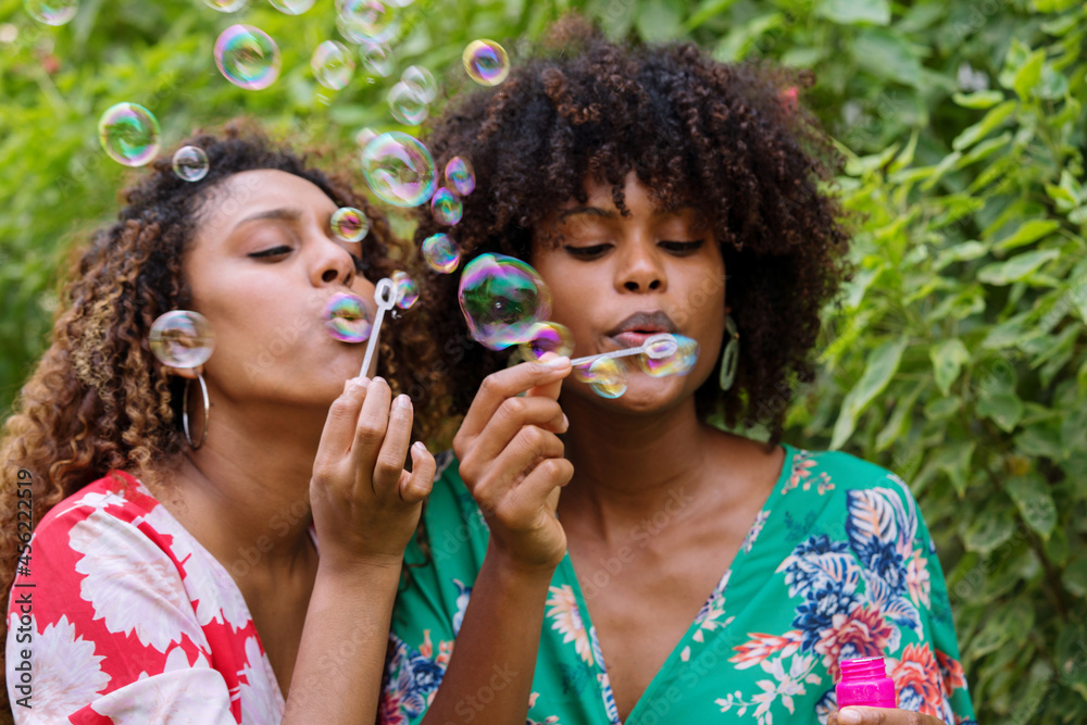 Two Happy Black woman with curly hair having fun blowing soap bubbles outdoors in the garden