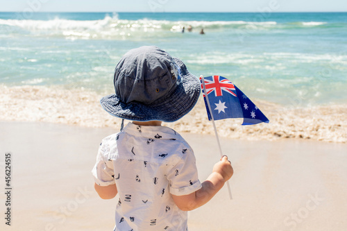 Little boy wearing hat holding Australian flag on a sandy ocean beach. Australia Day concept photo