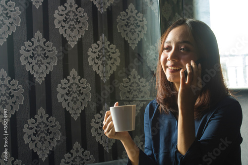 Relaxed Smilling Asian Woman drinking coffee while using a cell phone for work