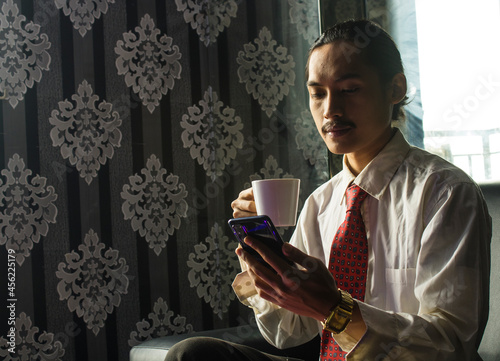 Relaxed Smilling Asian Man drinking coffee while using a cell phone for work