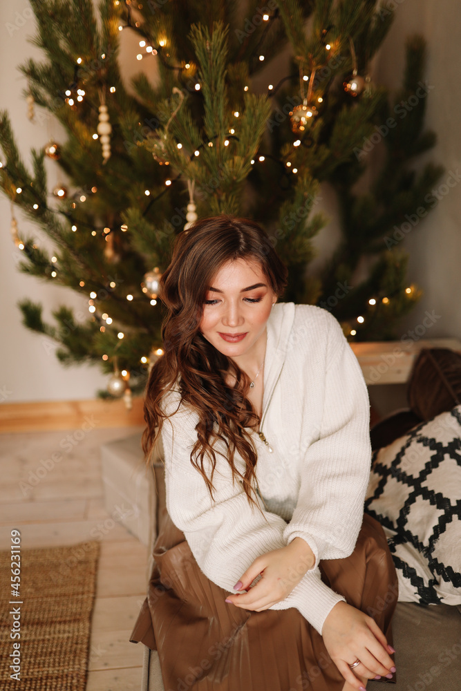 A sweet happy young woman in a cozy sweater is resting looking at a gift box sitting on the sofa by the Christmas tree during the winter holidays at home
