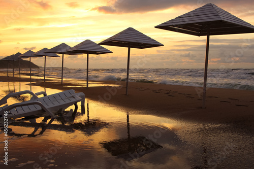 Umbrellas and sun loungers on wet sand by the beach in stormy weather in sunset light and sun reflections 