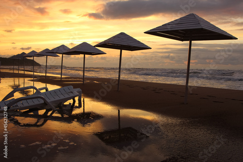 umbrellas and sun lounger on wet sand by the beach in stormy weather in sunset light and sun reflections 