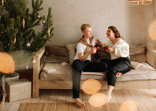 Happy lovers a man and a woman in cozy sweaters embrace spend time together celebrate Christmas relaxing on the sofa by the Christmas tree in the holidays in the interior in the style of eco friendly © Елизавета Старкова