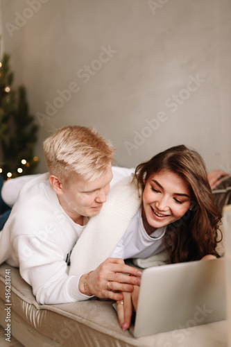 A smiling young man and a woman in love are sitting on the sofa hugging working using wireless technologies looking at a laptop and a phone during the winter holidays in a cozy house
