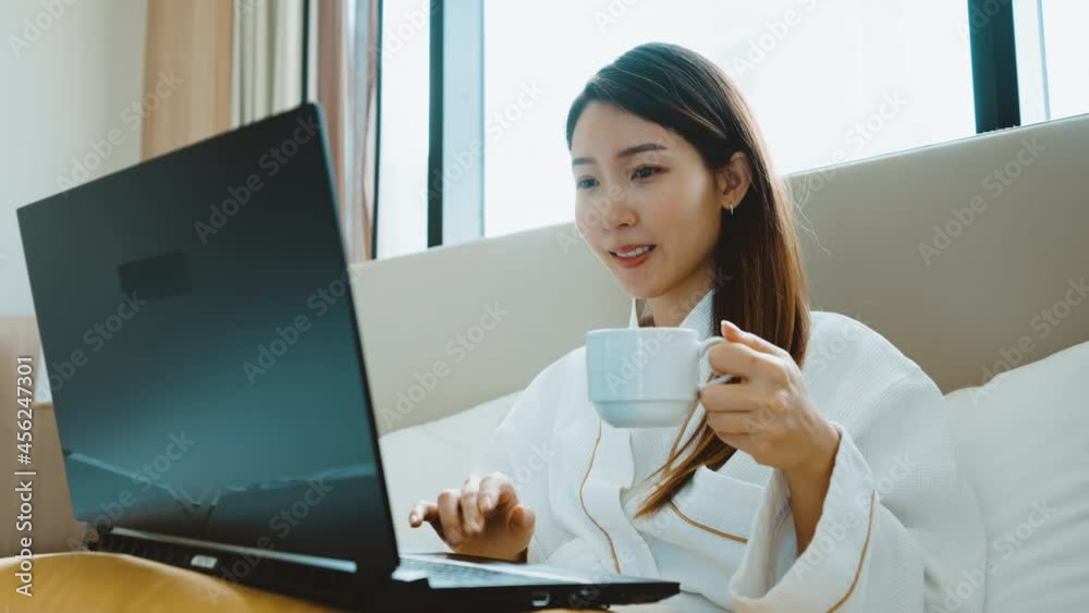 Portrait of Asian woman wearing bathrobe using a laptop for video call while have a morning coffee on bed in bedroom Portrait of Asian woman wearing bathrobe using a laptop for video call while have a morning coffee on bed in bedroom