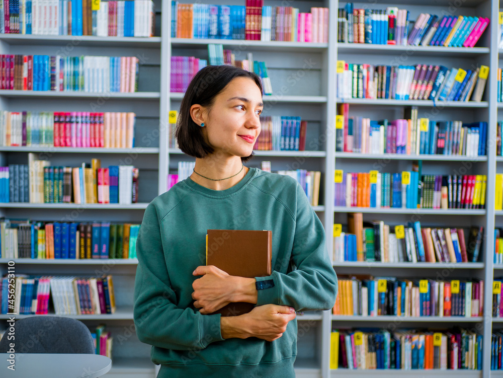 Young attractive female student reads a book preparing for an exam while against the backdrop of bookshelves. Young attractive female student reads a book preparing for an exam while against the backdrop of bookshelves.