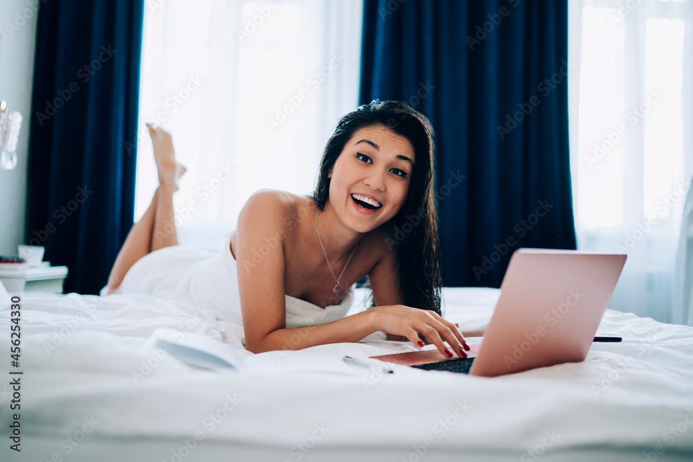 Overjoyed influence blogger with digital netbook computer lying at comfortable bed in hotel room and smiling at camera during morning time for creating content, happy Asian freelancer with laptop Overjoyed influence blogger with digital netbook computer lying at comfortable bed in hotel room and smiling at camera during morning time for creating content, happy Asian freelancer with laptop