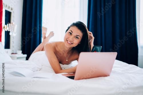 Portrait of happy Asian blogger with modern cellphone and laptop technology smiling at camera during leisure time in hotel bedroom, cheerful freelancer with digital smartphone and netbook resting