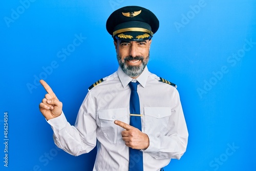 Middle age man with beard and grey hair wearing airplane pilot uniform smiling and looking at the camera pointing with two hands and fingers to the side.