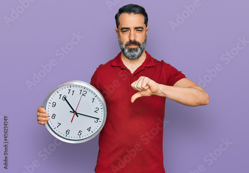 Middle aged man with beard holding big clock with angry face, negative sign showing dislike with thumbs down, rejection concept