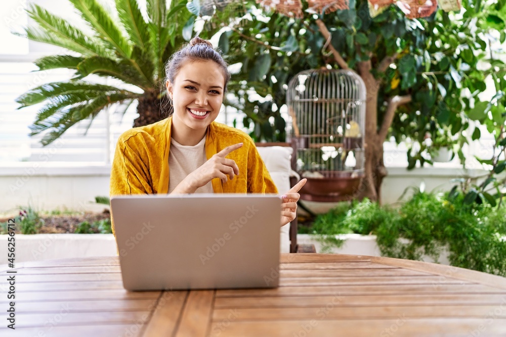 Beautiful young brunette woman using laptop sitting on the terrace at home smiling and looking at the camera pointing with two hands and fingers to the side. Beautiful young brunette woman using laptop sitting on the terrace at home smiling and looking at the camera pointing with two hands and fingers to the side.