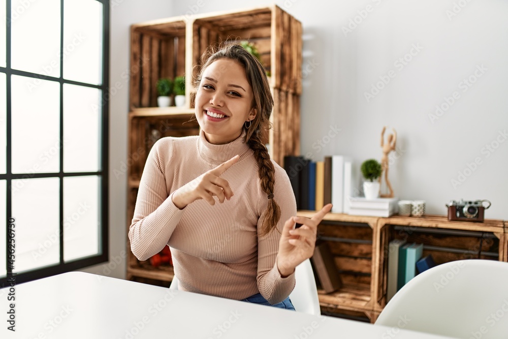 Young hispanic girl wearing casual clothes sitting on the table at home smiling and looking at the camera pointing with two hands and fingers to the side. Young hispanic girl wearing casual clothes sitting on the table at home smiling and looking at the camera pointing with two hands and fingers to the side.