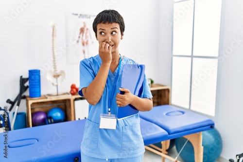 Young hispanic woman with short hair working at pain recovery clinic looking stressed and nervous with hands on mouth biting nails. anxiety problem.