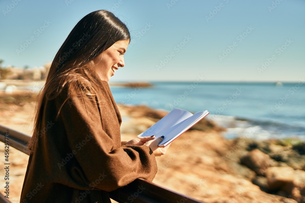 Young hispanic woman smiling happy reading book at the beach