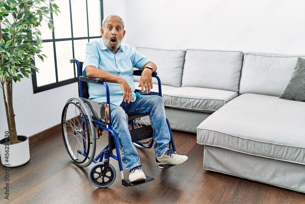 Handsome senior man sitting on wheelchair at the living room afraid and shocked with surprise expression, fear and excited face. Handsome senior man sitting on wheelchair at the living room afraid and shocked with surprise expression, fear and excited face.